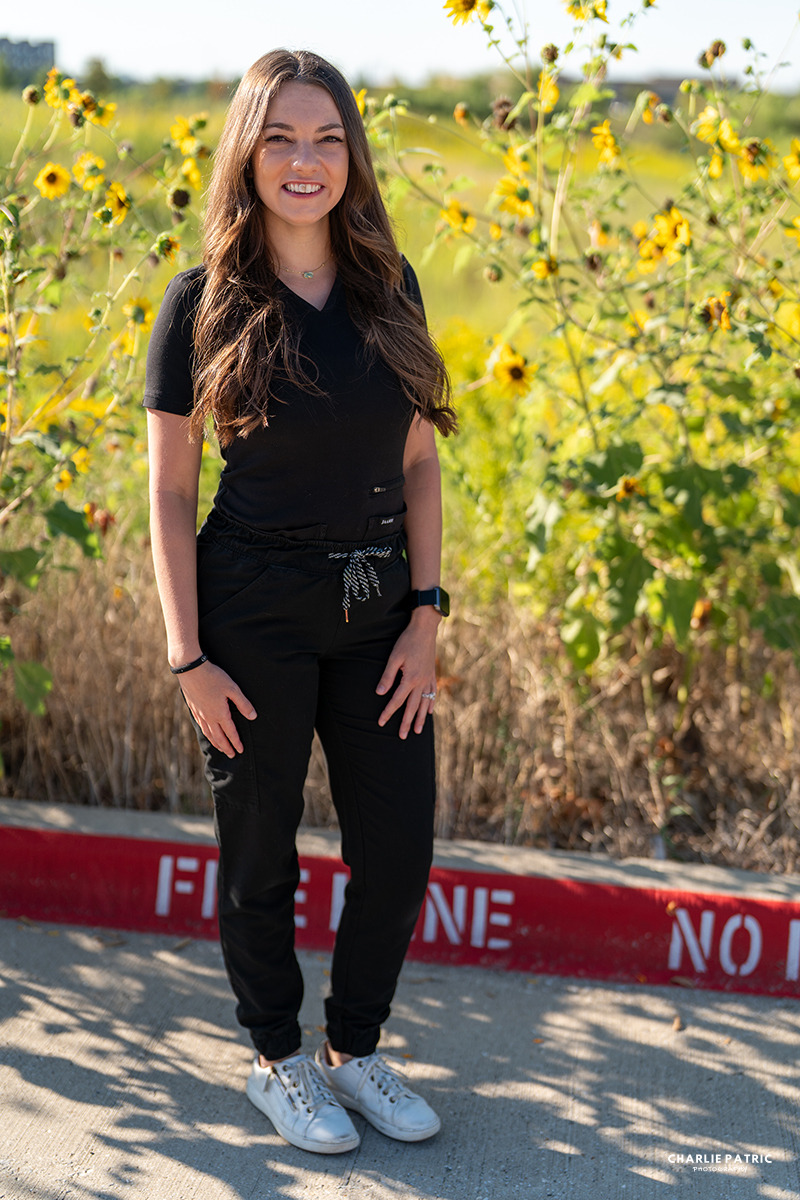 A woman with long brown hair, wearing a black outfit and white sneakers, stands on a Frisco sidewalk by yellow flowers and a red curb marked "FIRE LANE NO PARKING.