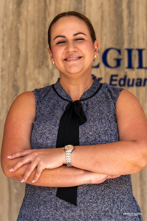 A woman with hair pulled back stands with arms crossed, smiling confidently in a sleeveless gray top with a black bow and a watch, embodying the perfect corporate image boost for professional headshots Frisco, against a beige wall with partial blue text.