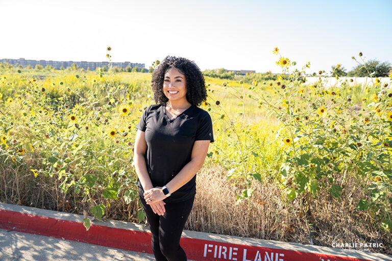 A woman in black scrubs stands smiling in front of sunflowers and tall grass near a curb labeled "FIRE LANE" on a sunny day, creating an inviting scene perfect for professional headshots or use as an online image.