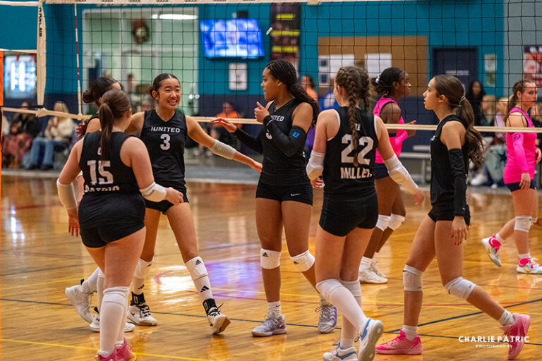 A girls’ volleyball team in black uniforms celebrates on an indoor court during a game, captured in dynamic sports photography for college recruitment, with another team in pink visible in the background.