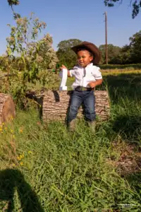 A young child in a cowboy hat, white shirt, jeans, and boots sits on a log holding a large number one in a grassy area with trees and wildflowers—perfect inspiration for photography venues or Frisco events.