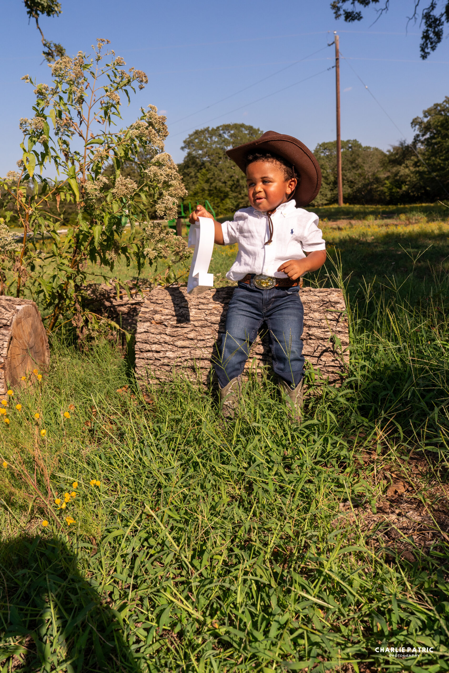 A young child in a cowboy hat, white shirt, jeans, and boots sits on a log holding a large number one in a grassy area with trees and wildflowers—perfect inspiration for photography venues or Frisco events.