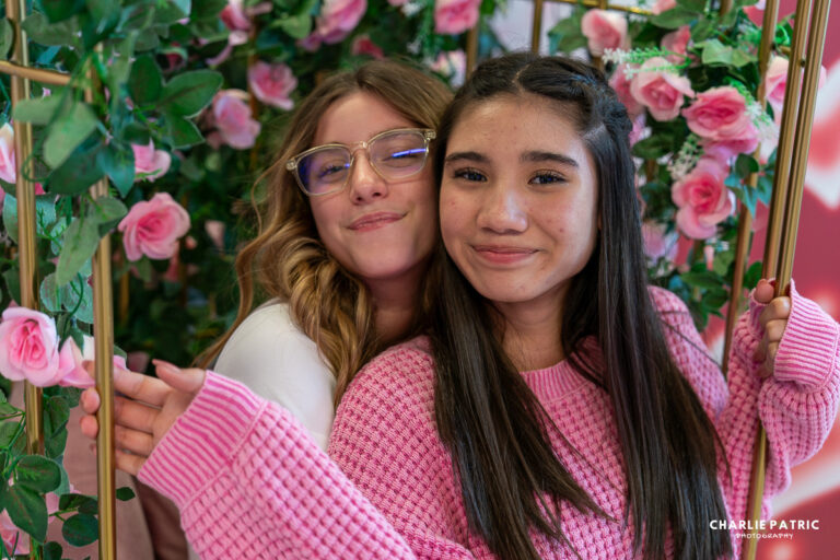 Two teenage girls smile and pose together on a swing decorated with pink roses and greenery, capturing moments of friendship. One wears glasses and a white sweater; the other, a pink sweater—perfect for local photography inspiration.