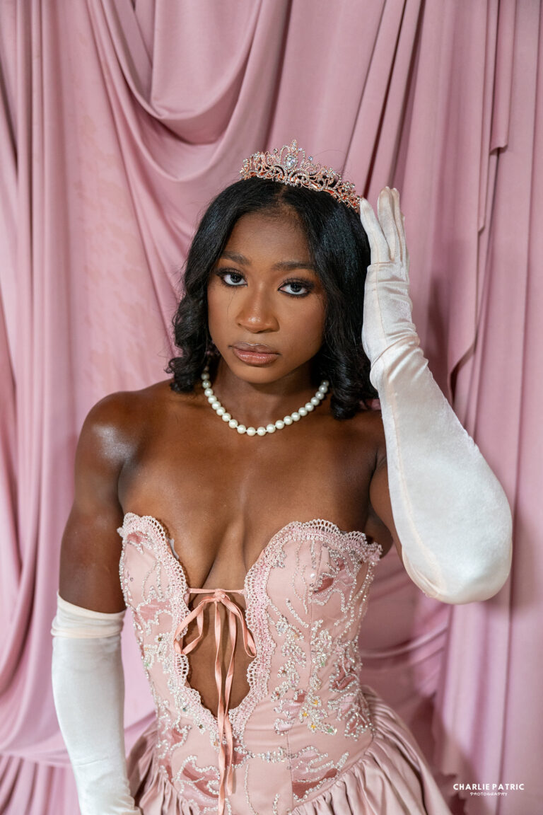 A woman wearing a pink lace corset gown, pearl necklace, long white gloves, and a tiara poses in front of pink draped fabric—an elegant style perfect for Frisco graduation photography services.