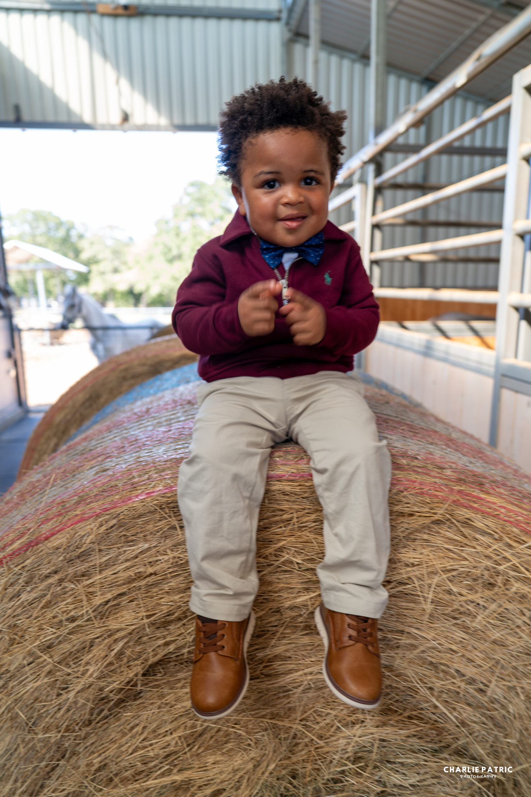 A young child in a maroon sweater, beige pants, and brown shoes sits on a large bale of hay inside a barn, capturing one of those memorable moments that our photography services are dedicated to preserving.