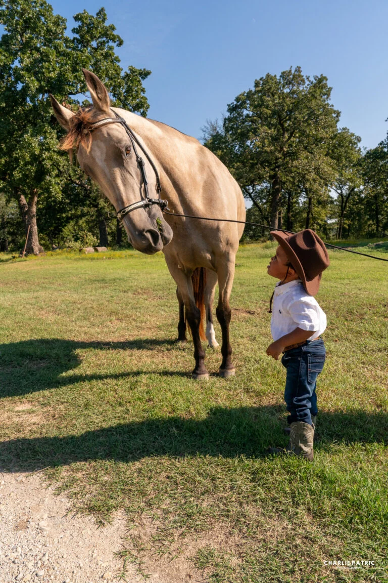 A young child in a cowboy hat and boots stands in front of a light brown horse on a grassy area with trees in the background, capturing a charming moment perfect for those seeking photography tips or enhancing experience in event photography.