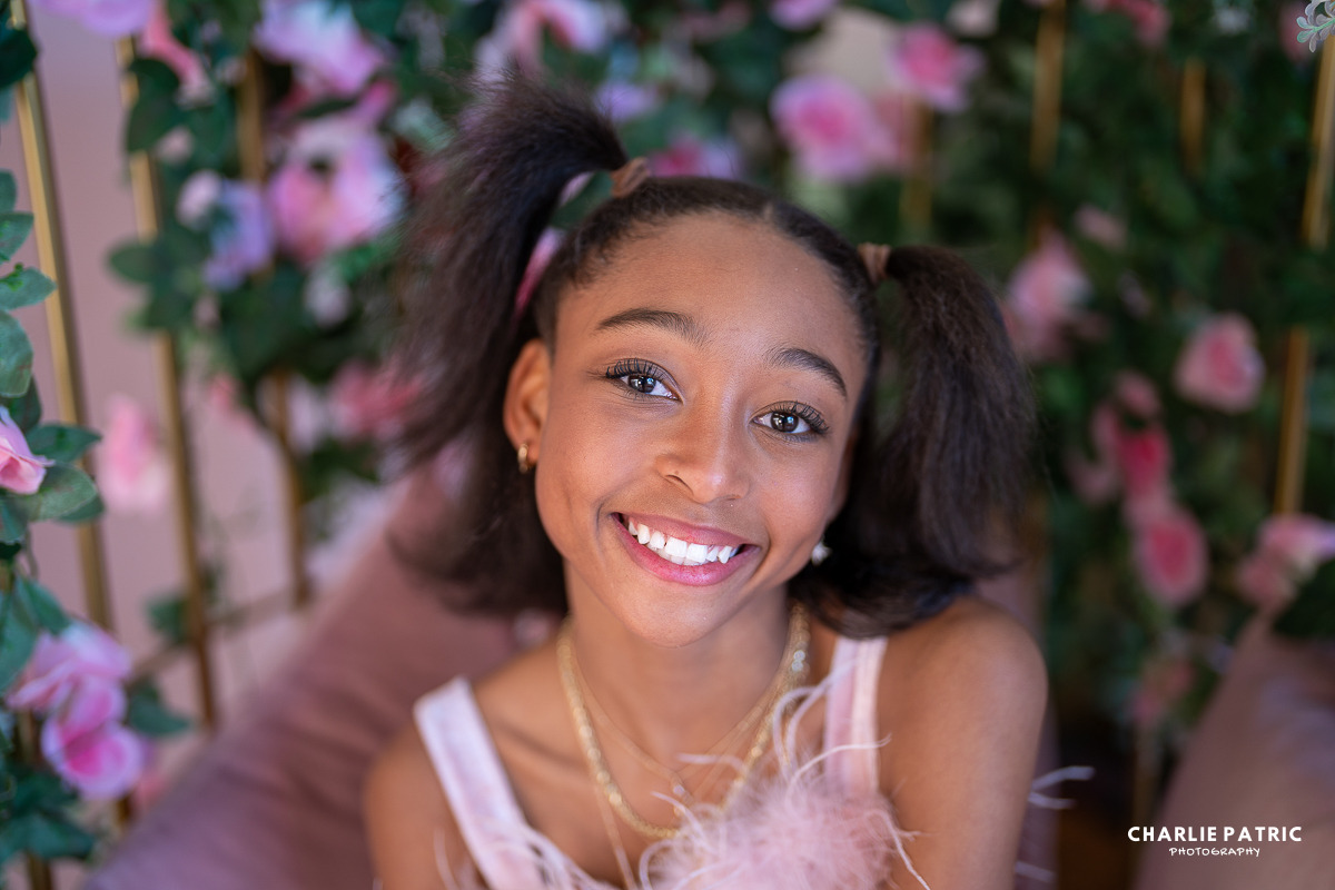 Young girl with pigtails smiling at the camera, seated in front of a floral background with pink flowers and greenery—perfect inspiration for Frisco Commons Park photography or anyone seeking creative photography tips.
