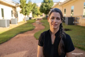 A woman with long gray-streaked hair in a braid stands outside on a gravel path between buildings, wearing a black shirt and smiling at the camera—perfect for corporate headshots Frisco or your next headshot update signs.