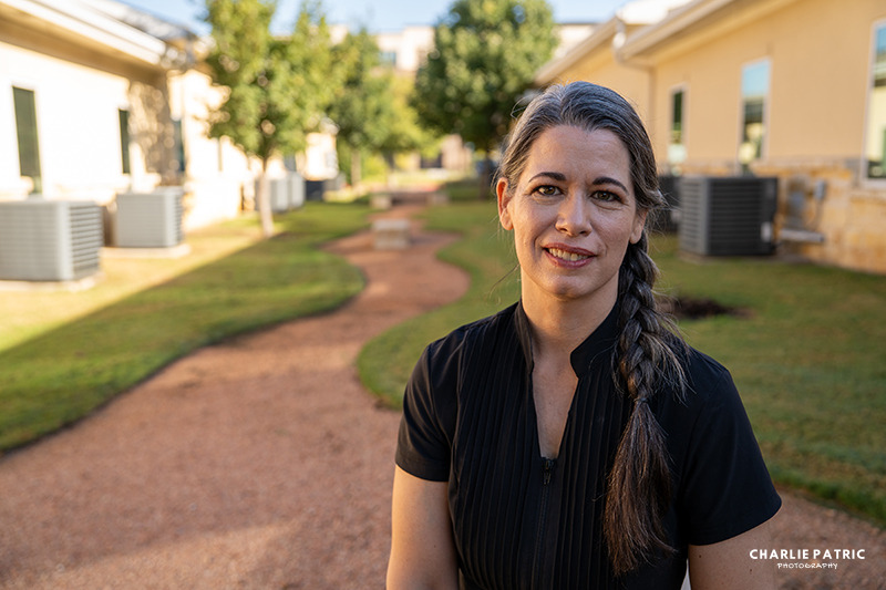A woman with long gray-streaked hair in a braid stands outside on a gravel path between buildings, wearing a black shirt and smiling at the camera—perfect for corporate headshots Frisco or your next headshot update signs.