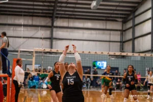 A volleyball player in a black jersey prepares to set the ball during an indoor match, with youth sports photography services capturing teammates and opponents watching intently.