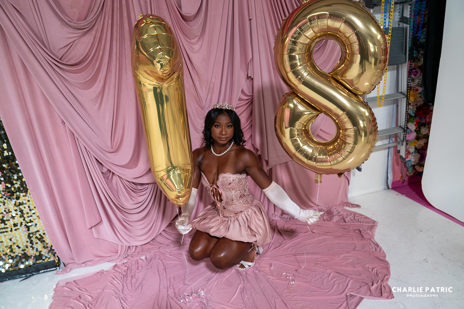 A young woman wearing a tiara and pink dress kneels on pink draped fabric, holding gold balloon numbers "1" and "8" against a pink backdrop, celebrating her 18th birthday with style fit for Frisco graduation photography services.