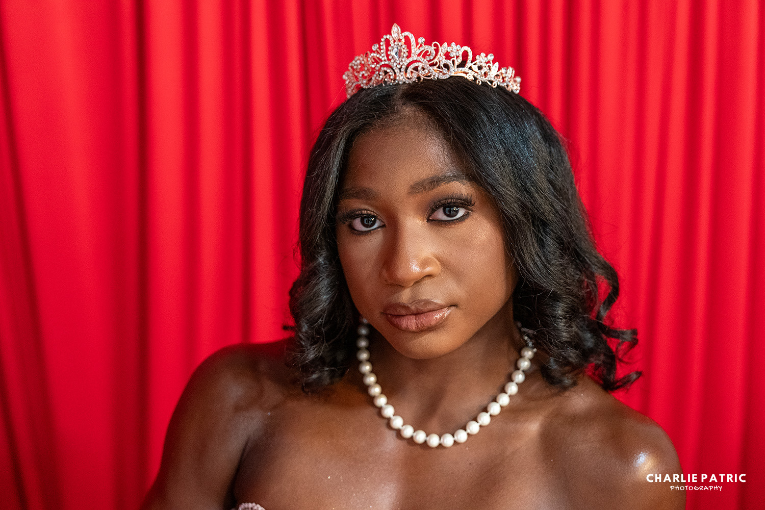 A woman wearing a tiara and pearl necklace stands in front of red curtains, looking directly at the camera—a perfect example of elegance captured by Frisco graduation photography services.