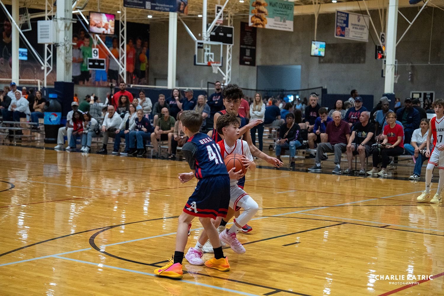 A youth basketball game in progress, with one player dribbling the ball while being defended by two opponents; spectators watch from the bleachers, capturing the excitement and energy that youth sports photography services aim to preserve.