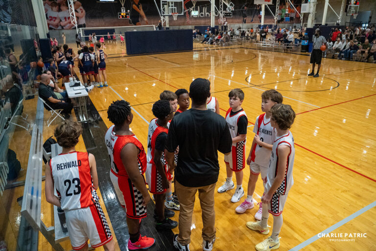 A youth basketball coach gives instructions to his team during a time-out; players in white and red uniforms stand in a huddle on a gym court, captured by professional youth sports photography services.