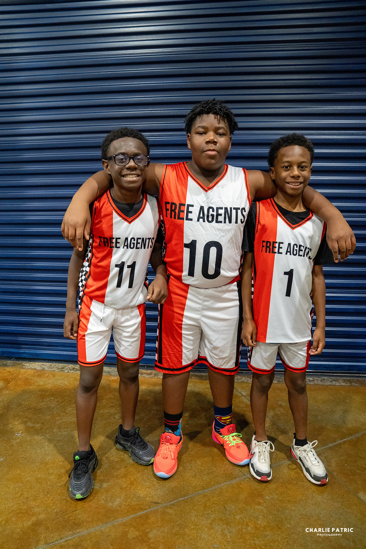 Three boys in “Free Agents” basketball uniforms stand together, smiling, with their arms around each other in front of a blue corrugated wall—captured by a Frisco sports event photographer.