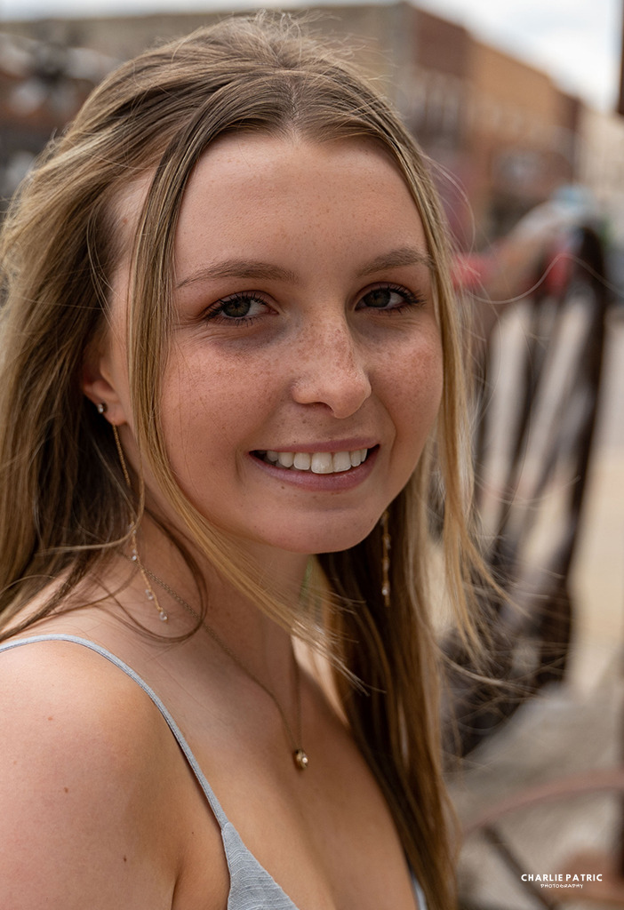 Young woman with long hair smiling outdoors, wearing a light-colored top and delicate jewelry, with blurred buildings in the background—capturing a joyful moment that highlights the style and quality of Frisco graduation photography services.