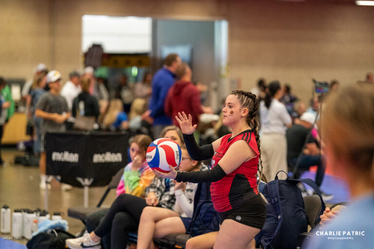 A young volleyball player in a red jersey holds a ball, preparing to serve, while others sit and watch in a busy indoor gym setting—perfectly captured by a Frisco sports event photographer.