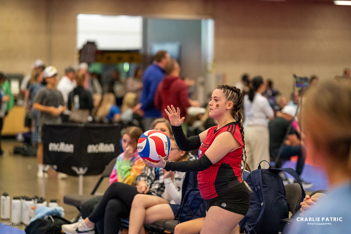 A young volleyball player in a red jersey holds a ball, preparing to serve, while others sit and watch in a busy indoor gym setting—perfectly captured by a Frisco sports event photographer.
