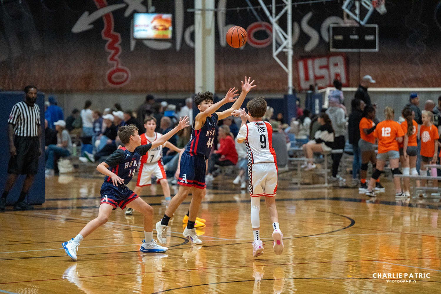 A youth basketball player in a white jersey shoots the ball while two defenders in dark jerseys jump to block him during an indoor game, perfectly captured by a skilled Frisco sports event photographer.