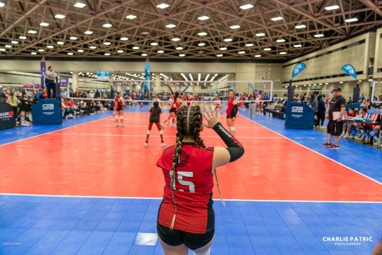 A volleyball player wearing a red jersey with number 15 prepares to serve during an indoor tournament, captured by a Frisco sports event photographer, with teams and spectators visible in the background.